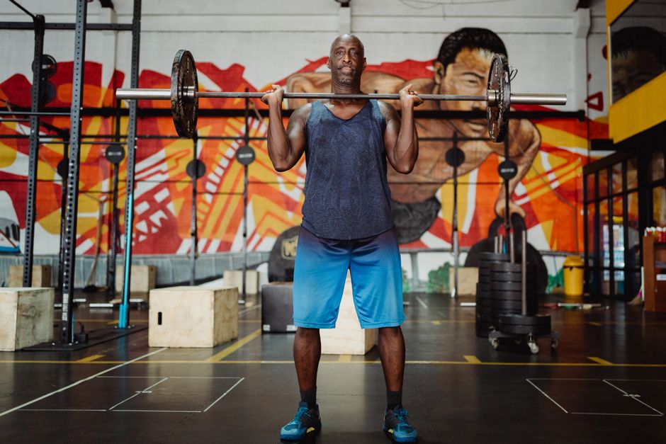 Side view of a man working out with a dumbbell and weighted vest in a gym setting.