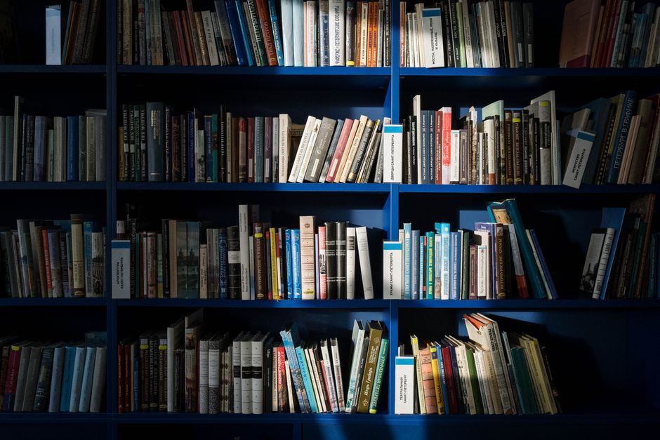 A person lying down, holding and reading George Orwell's 1984, surrounded by scattered books.