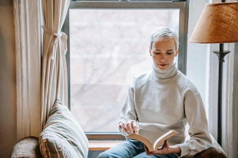 Focused pleasant female in casual outfit studying interesting novel on couch with cushion near window