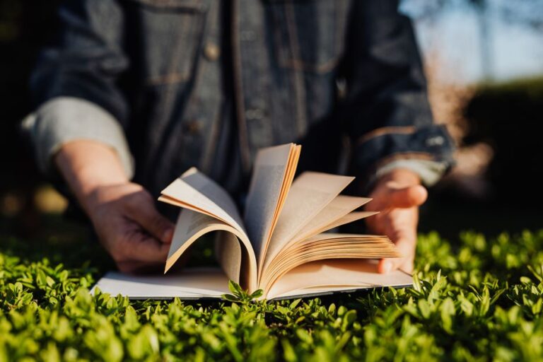 Crop unrecognizable male in casual clothes reading novel while standing at grassy surface on sunny summer day