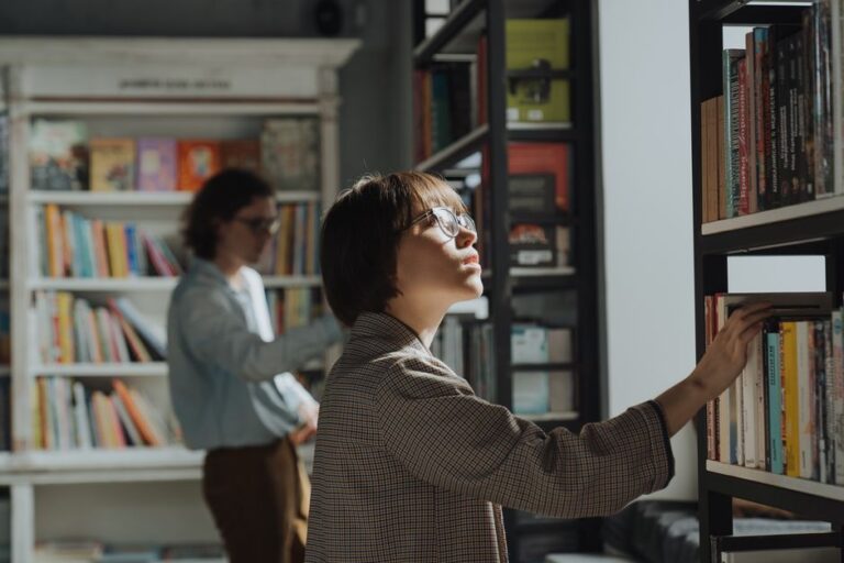 A young woman wearing glasses browses books in a well-lit library, symbolizing education and self-improvement.