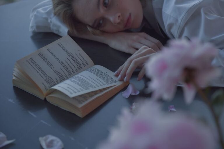 Caucasian woman in a contemplative mood, reading a book next to soft pink petals indoors.