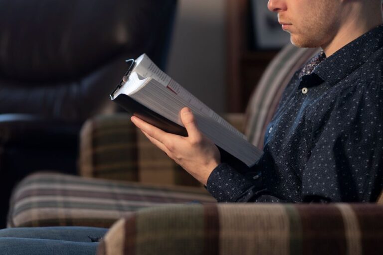 Adult male reading a Bible while seated on a comfortable chair indoors.